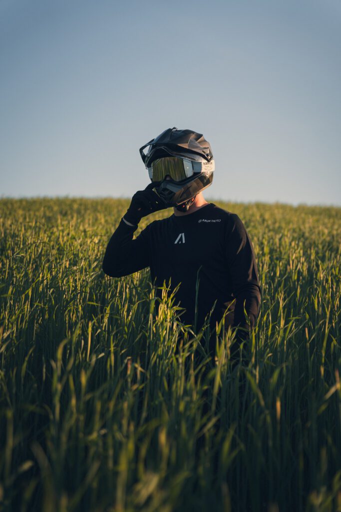 Adventure motorcyclist in a black riding shirt and helmet standing in a grassy field at sunset.