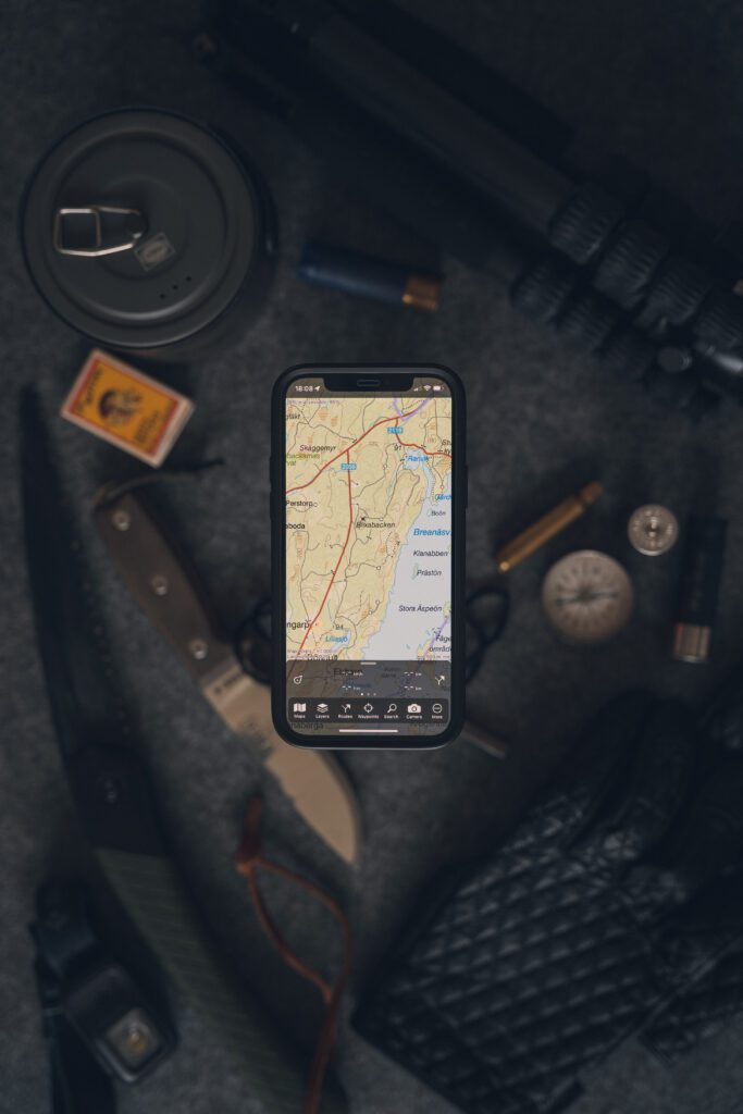 Smartphone displaying a detailed map, surrounded by camping and navigation essentials like a knife, compass, matches, and a cooking kit