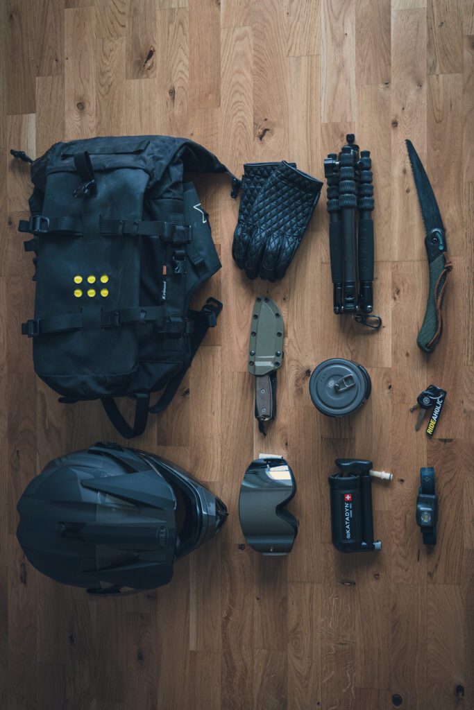 Flat lay of essential motorcycle camping gear including a black saddlebag, gloves, helmet, knife, folding saw, goggles, compact water filter, cooking kit, headlamp, and a small tripod.