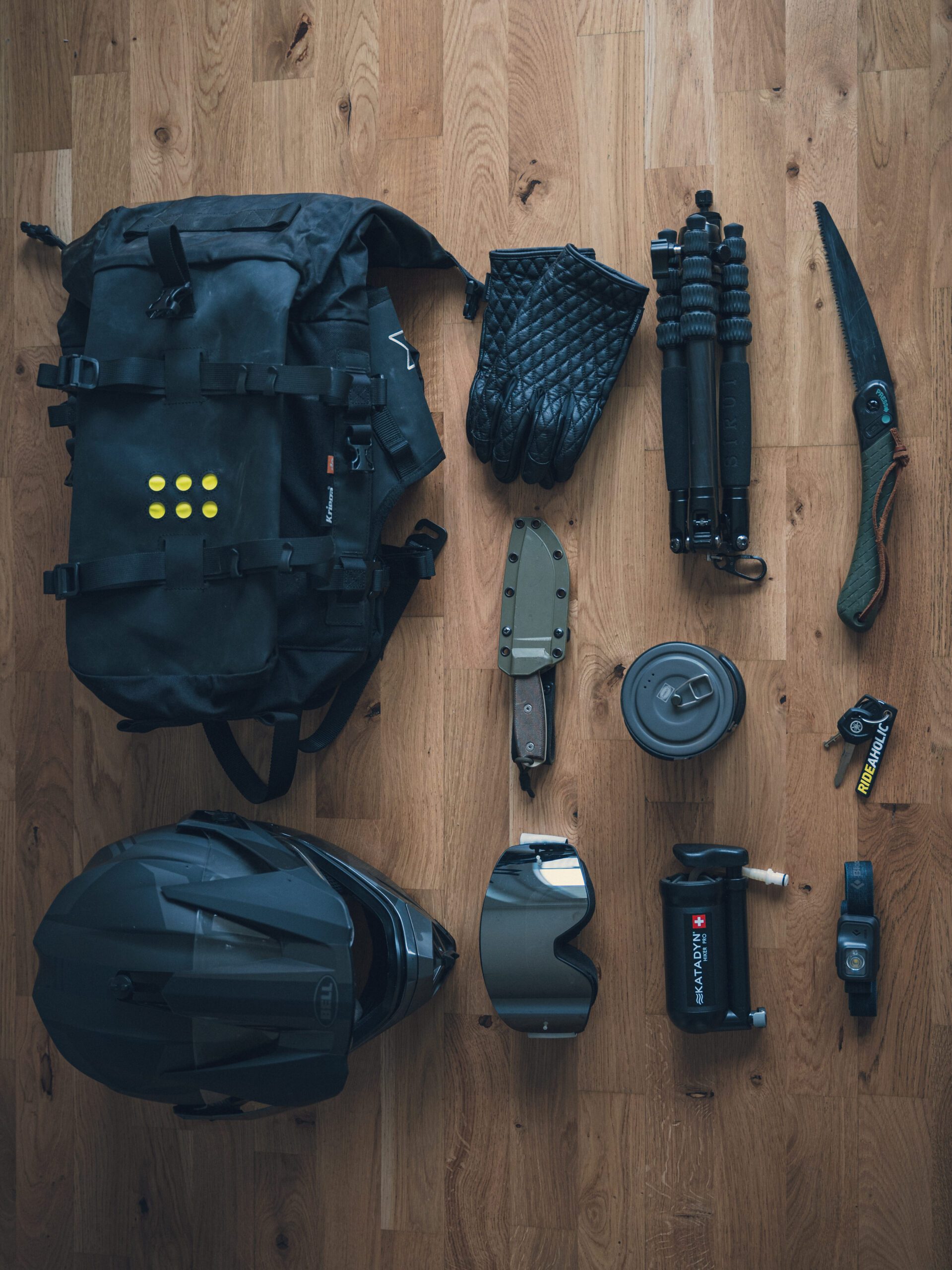 Flat lay of essential motorcycle camping gear including a black saddlebag, gloves, helmet, knife, folding saw, goggles, compact water filter, cooking kit, headlamp, and a small tripod.